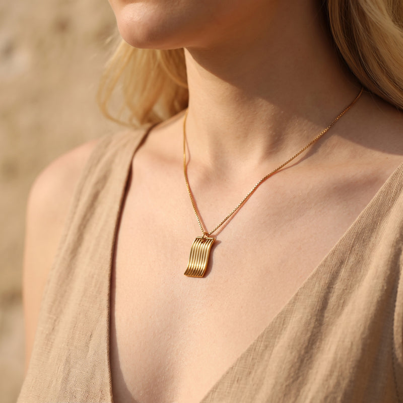 Gold-tone fluid wave pendant necklace on a box chain worn on a woman's neckline.