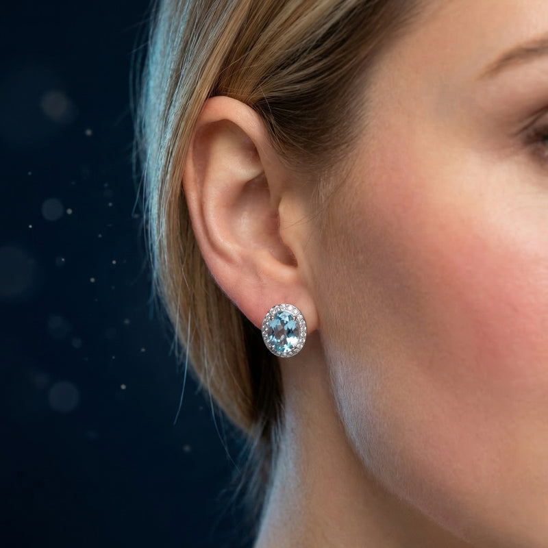 Close-up of a white gold oval blue topaz halo drop earring worn on a model's ear against a dark background.