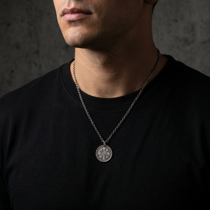 Angled close-up of a man wearing the silver coin pendant necklace over a black T-shirt against a concrete wall.