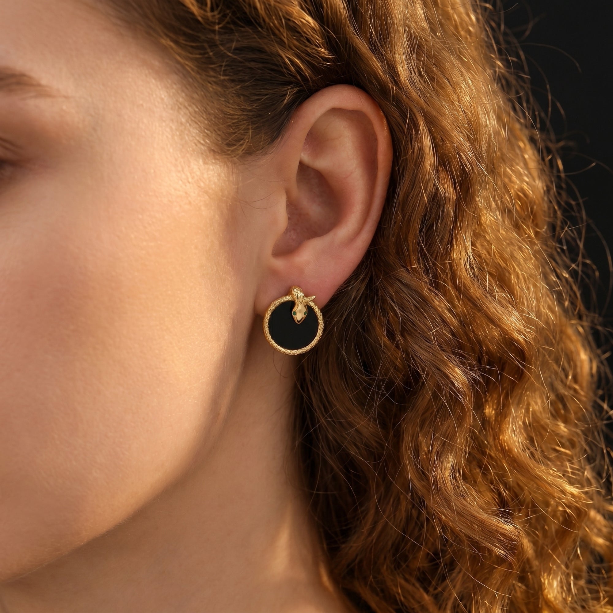 Model wearing a gold plated Serpens onyx earring featuring a snake head design with green eyes.