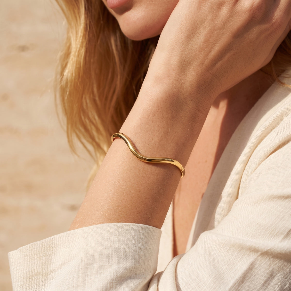 Gold-tone sculpted wave cuff bracelet worn on a model's wrist beside a light beige linen sleeve.