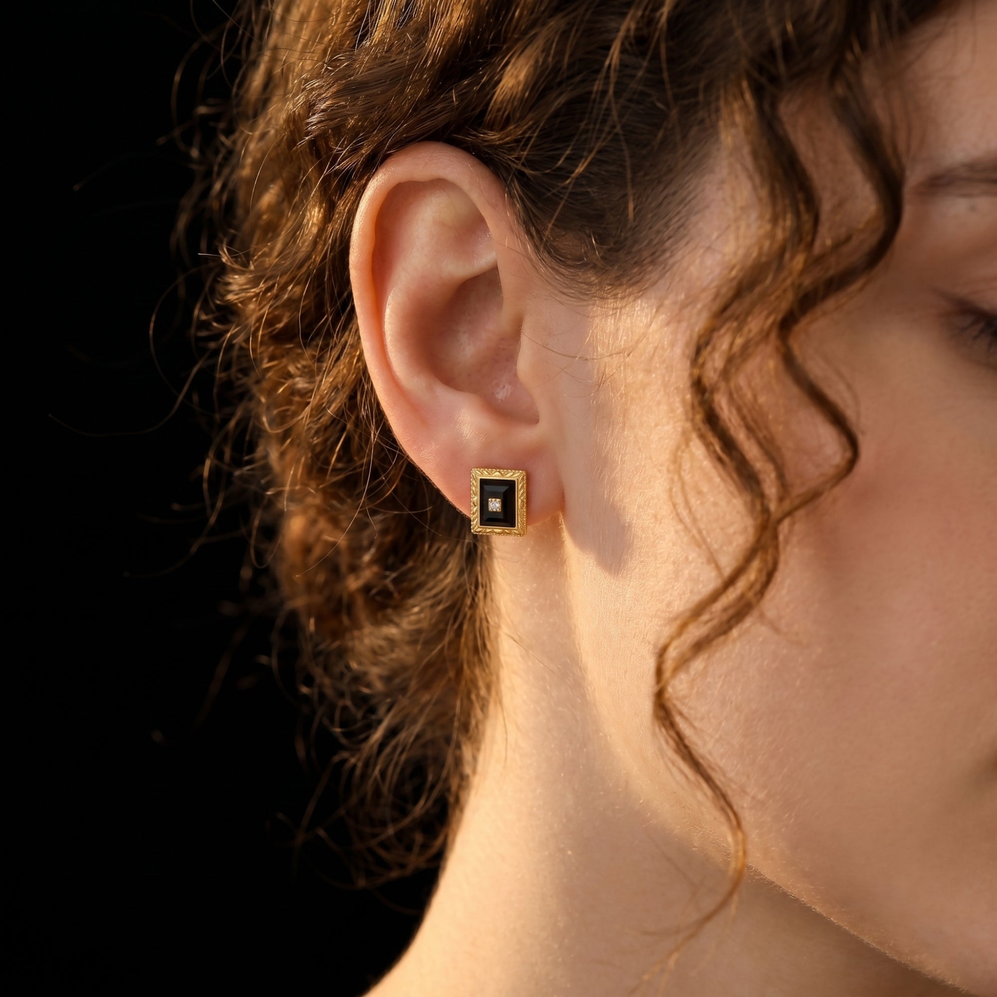Woman wearing a yellow gold square-cut black onyx stud earring with a center accent stone.