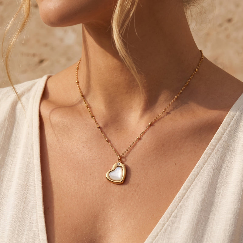 Model wearing a gold-tone heart necklace with an iridescent white insert and satellite chain over a white top.