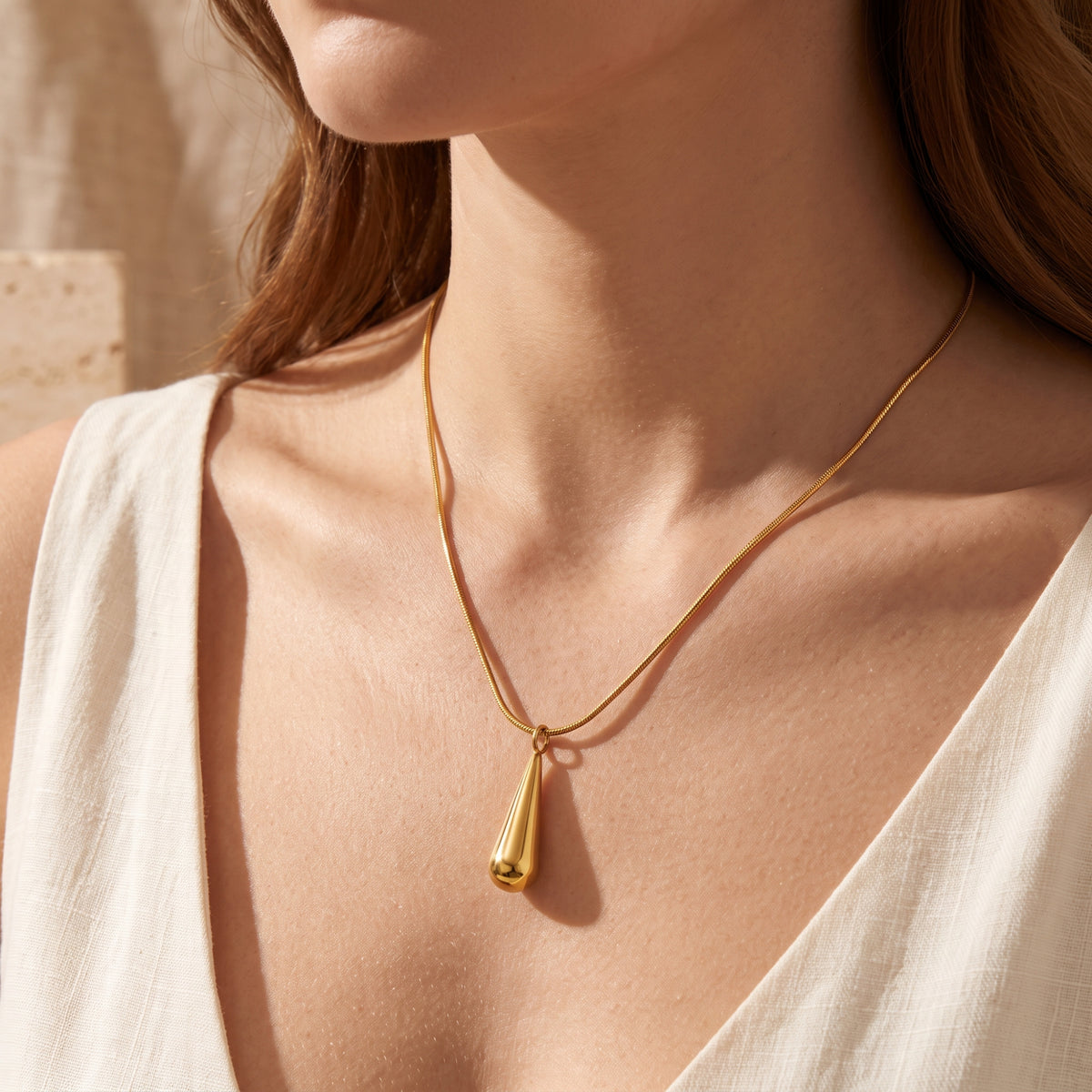 Model wearing a gold-tone teardrop pendant necklace with a snake chain and a light cream top.