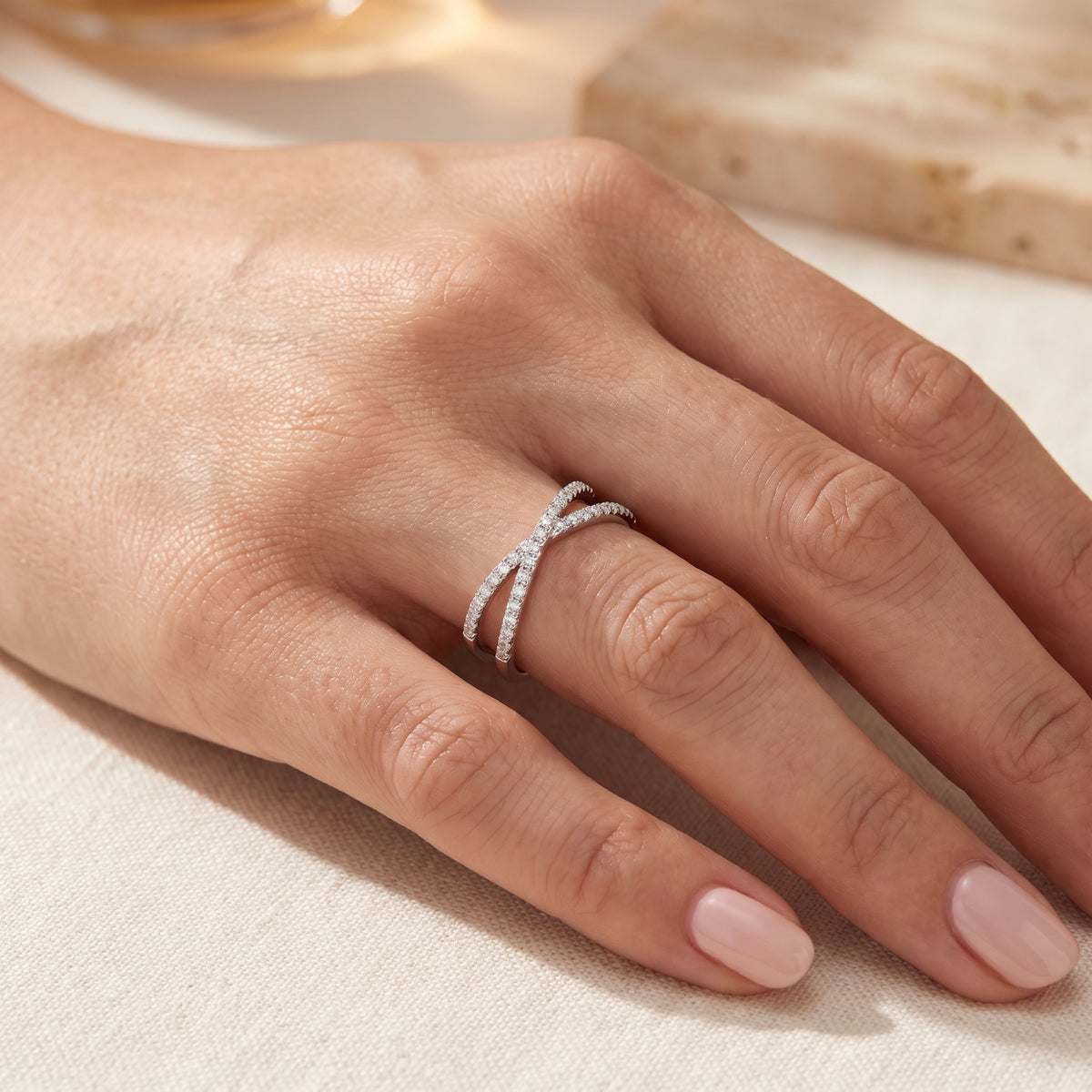 Silver crossover ring with small clear stones worn on a hand with nude manicured nails.