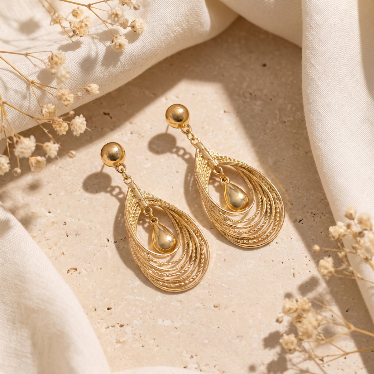 Both gold-toned layered teardrop earrings arranged on a stone surface among white fabric and dried flowers.