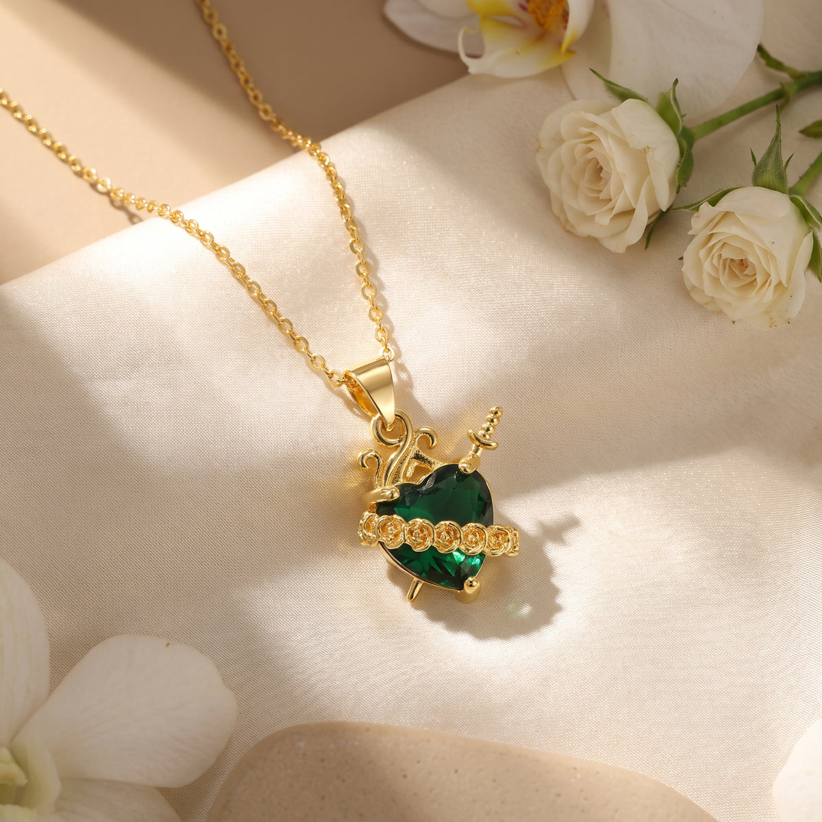 Gold-tone heart and dagger pendant necklace displayed on cream fabric alongside white roses.