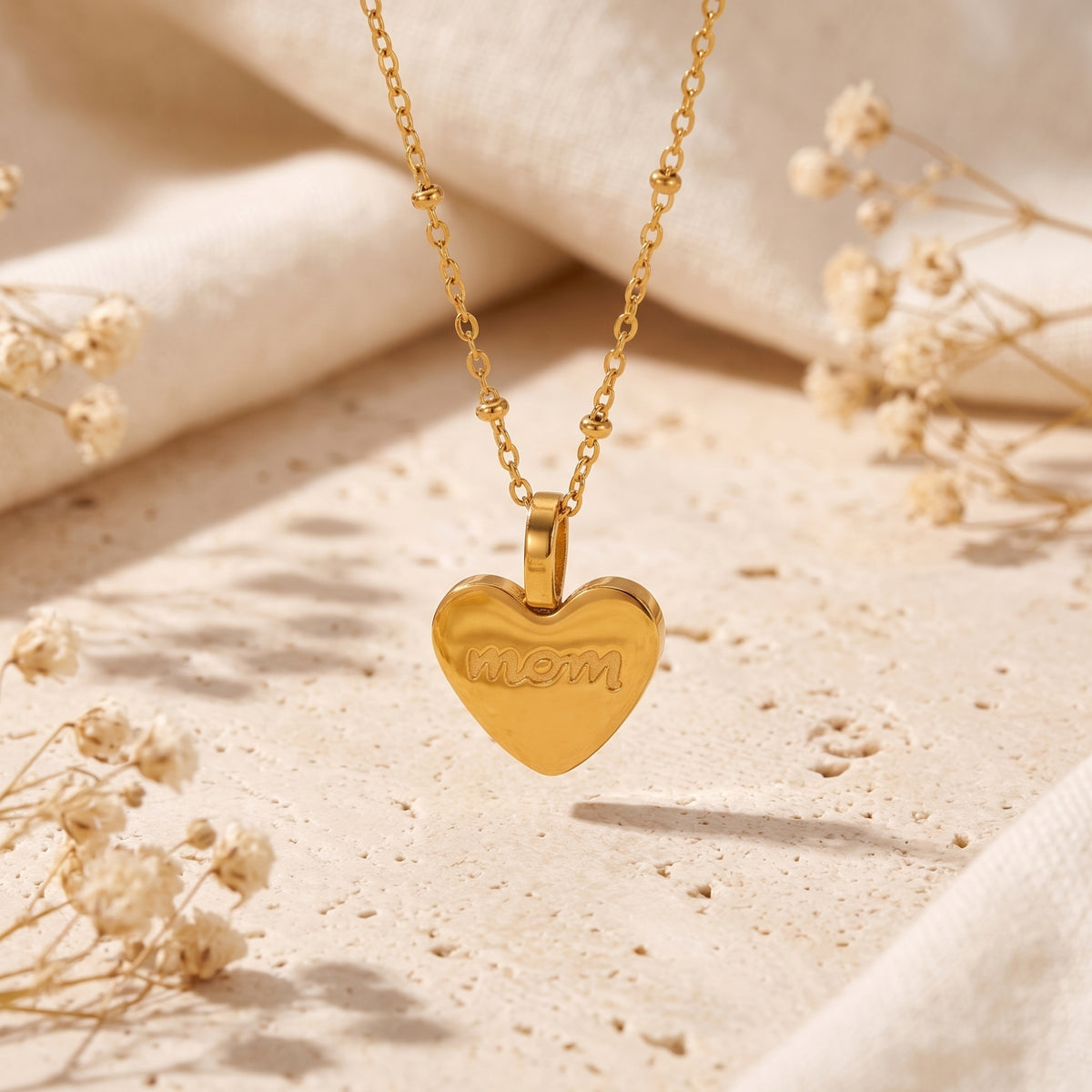 Front view of the gold-tone heart pendant engraved with 'mom', hanging over a beige stone surface.