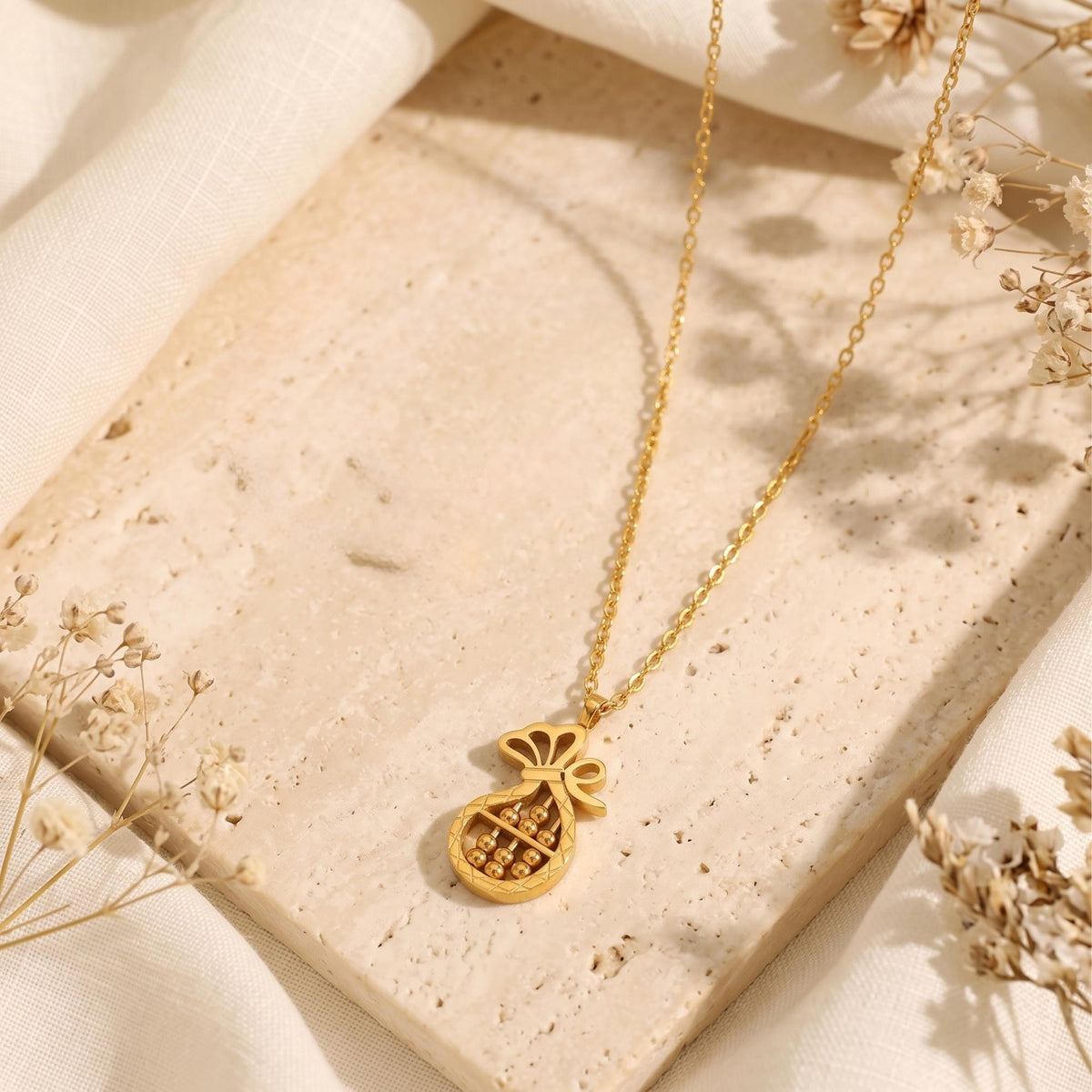 Gold-tone abacus money bag necklace displayed flat on travertine tile beside dried flowers.