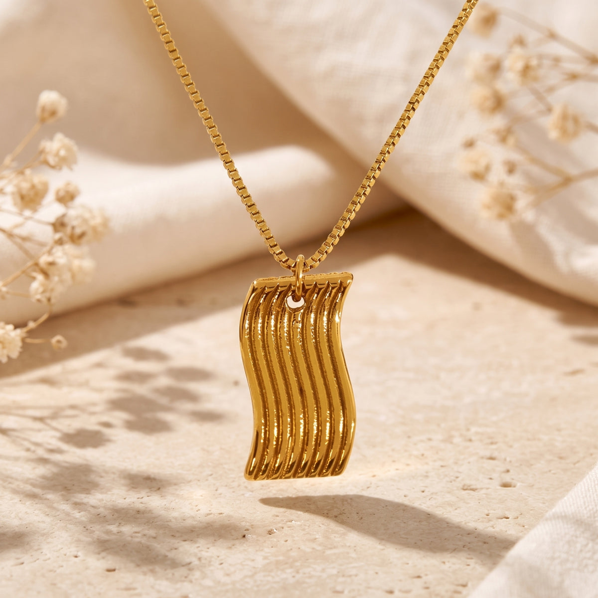 Gold-tone wavy rectangular pendant hanging on a box chain above a stone slab background.