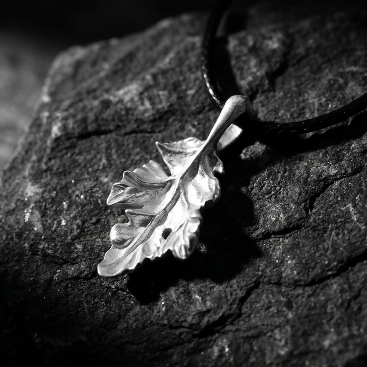 Angled close-up of a silver oak leaf pendant resting on dark rocks, showing sculpted leaf textures.