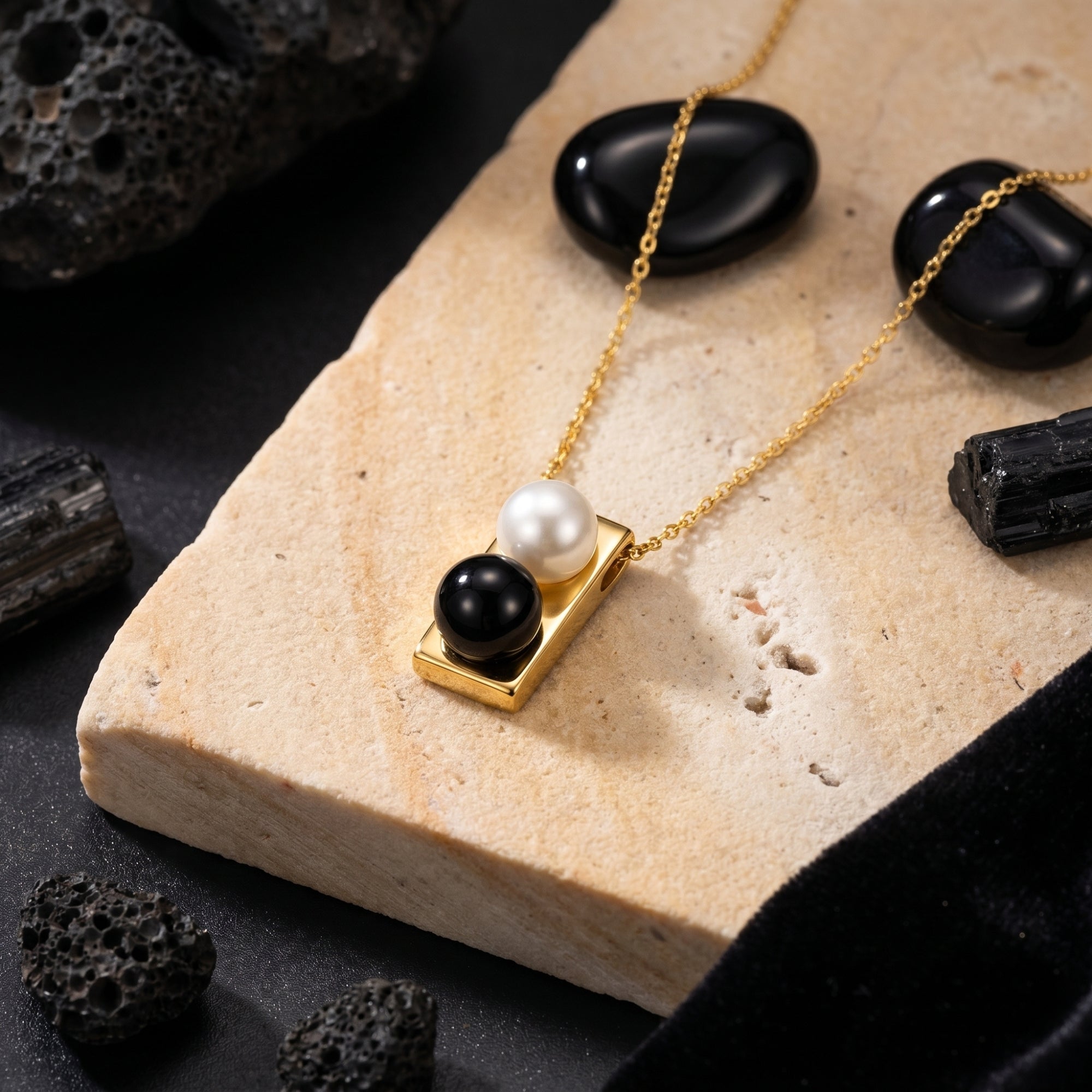 Gold pendant necklace with a round white pearl and black onyx stone resting on a light-colored rock.