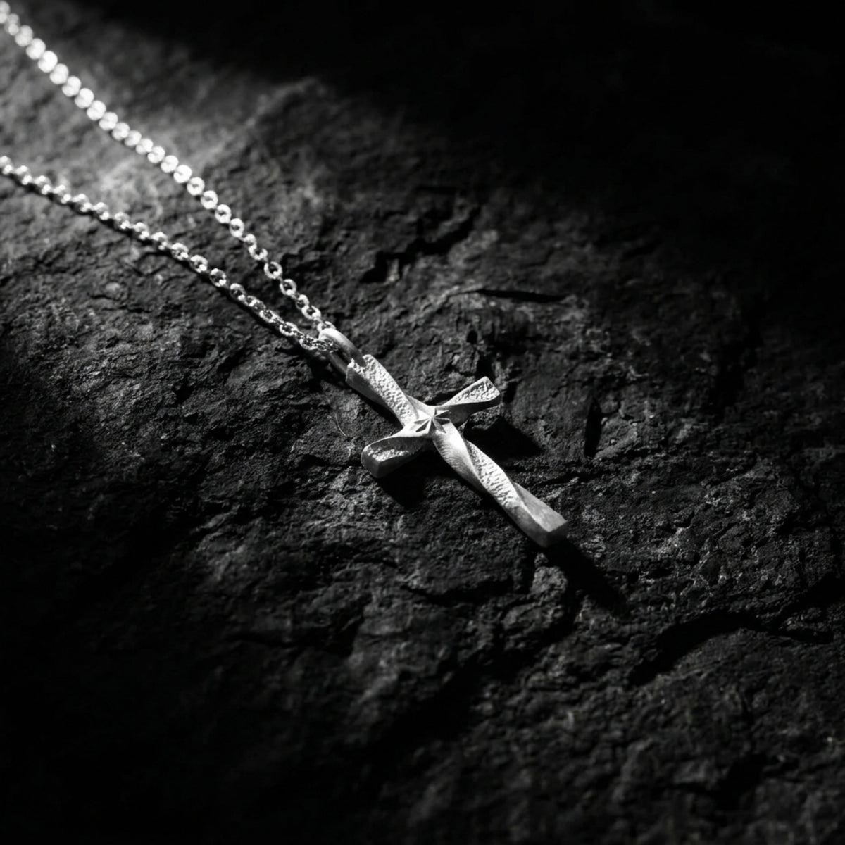 Silver twisted cross pendant resting diagonally on a dark textured rock surface under focused lighting.