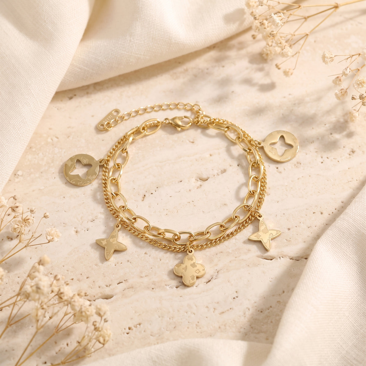 Full view of the gold-tone double-chain charm bracelet arranged flat on a beige stone slab with dried flowers.