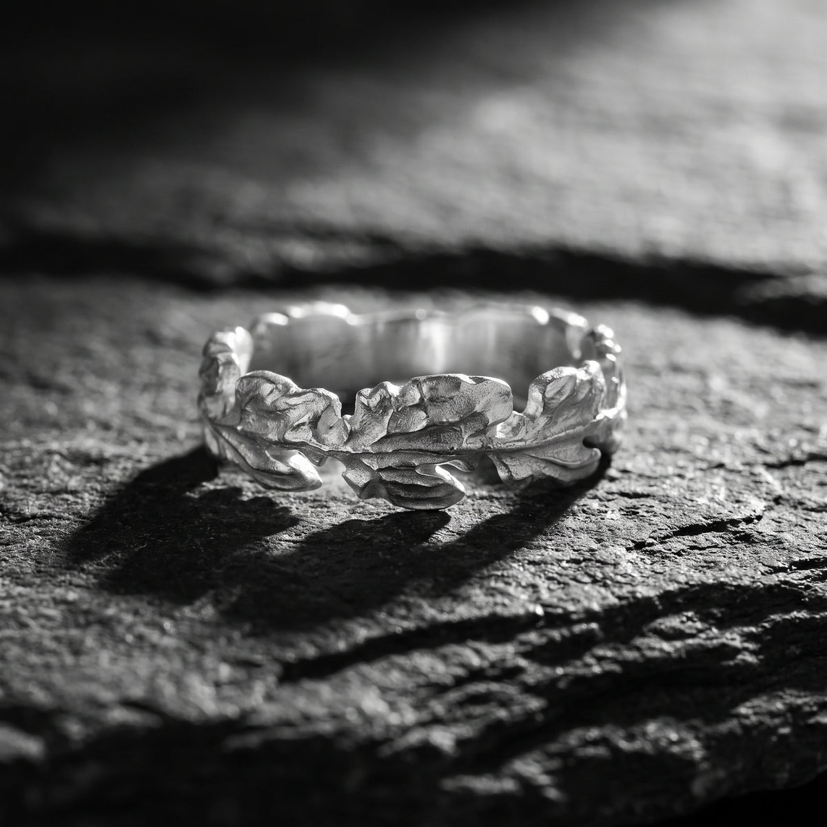 Close-up of a silver oak leaf ring resting horizontally on a rough dark textured surface.
