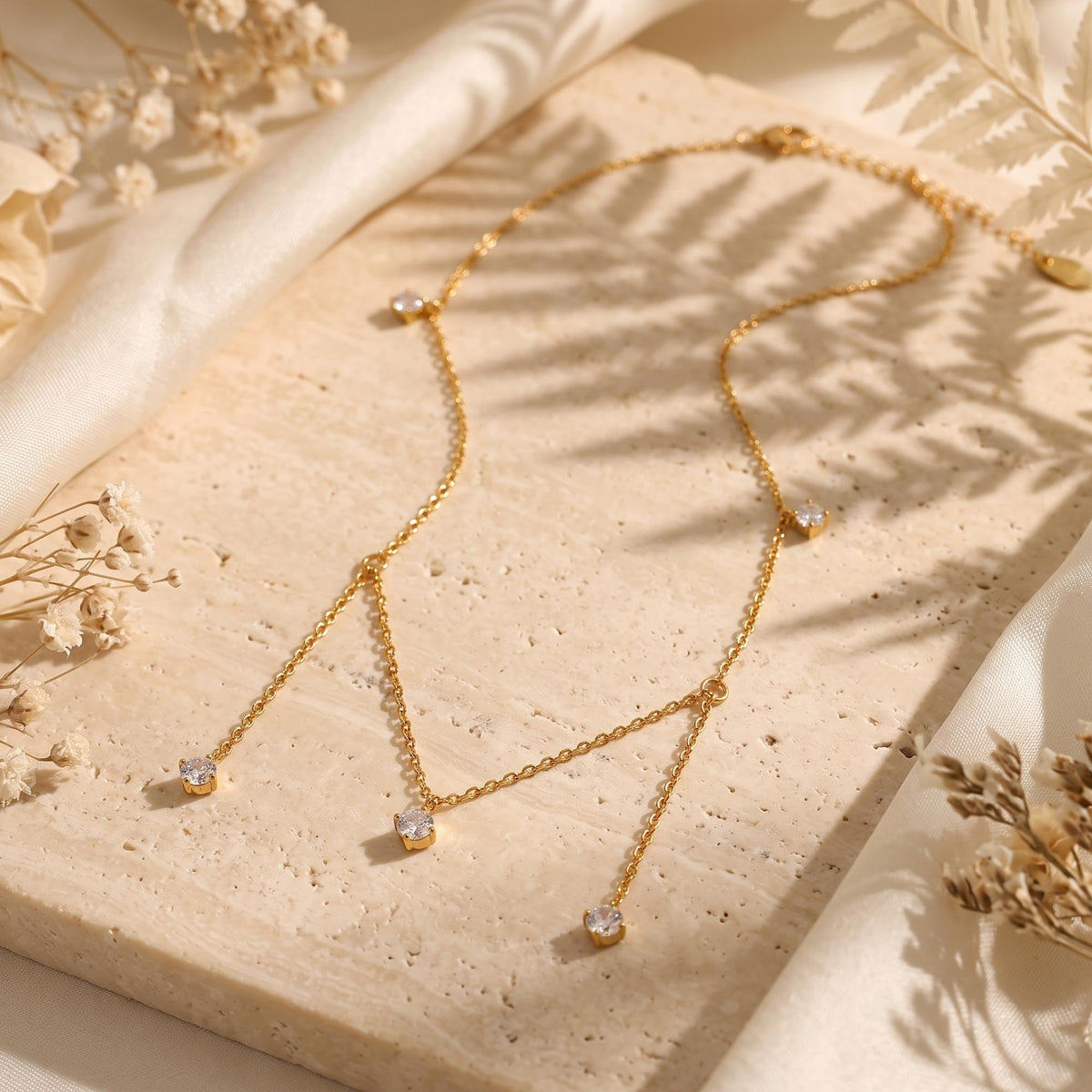Full flat lay of a gold-tone five-drop necklace with clear stones on a beige stone surface.
