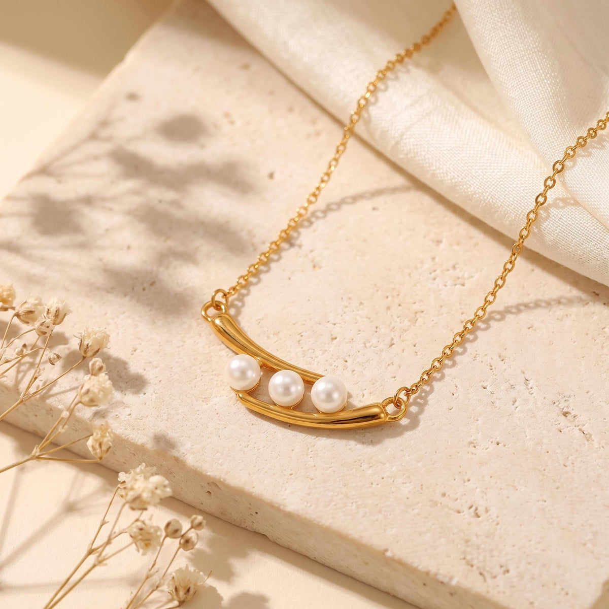 Flat lay of the gold-tone crescent necklace with three simulated pearls on a pale stone slab beside white fabric.