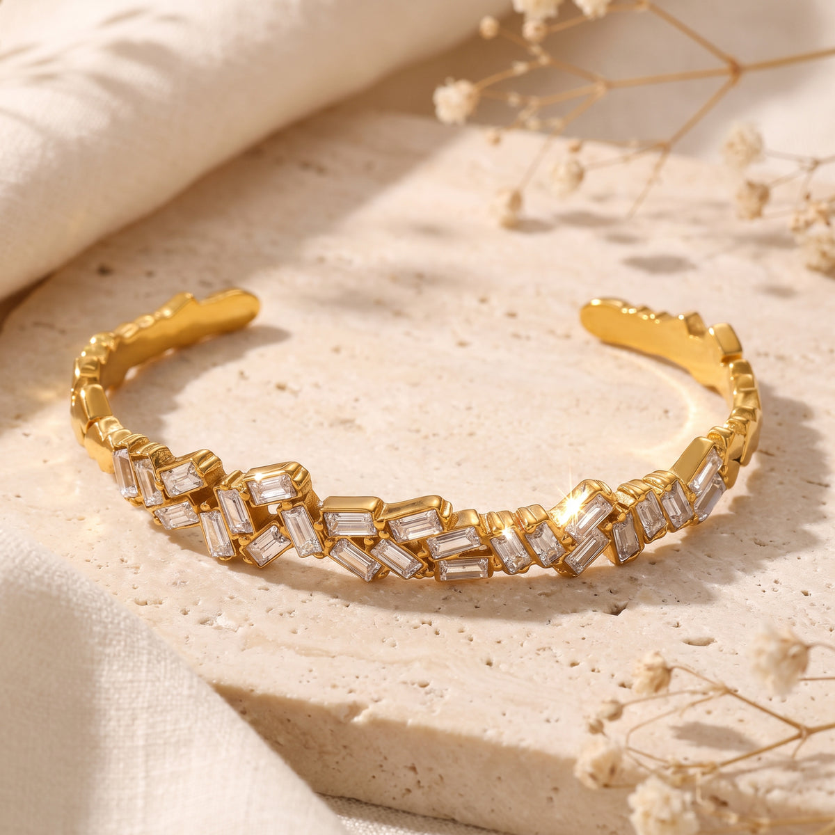 Gold-tone open cuff bracelet with clear baguette-cut stones, displayed flat on a travertine surface.
