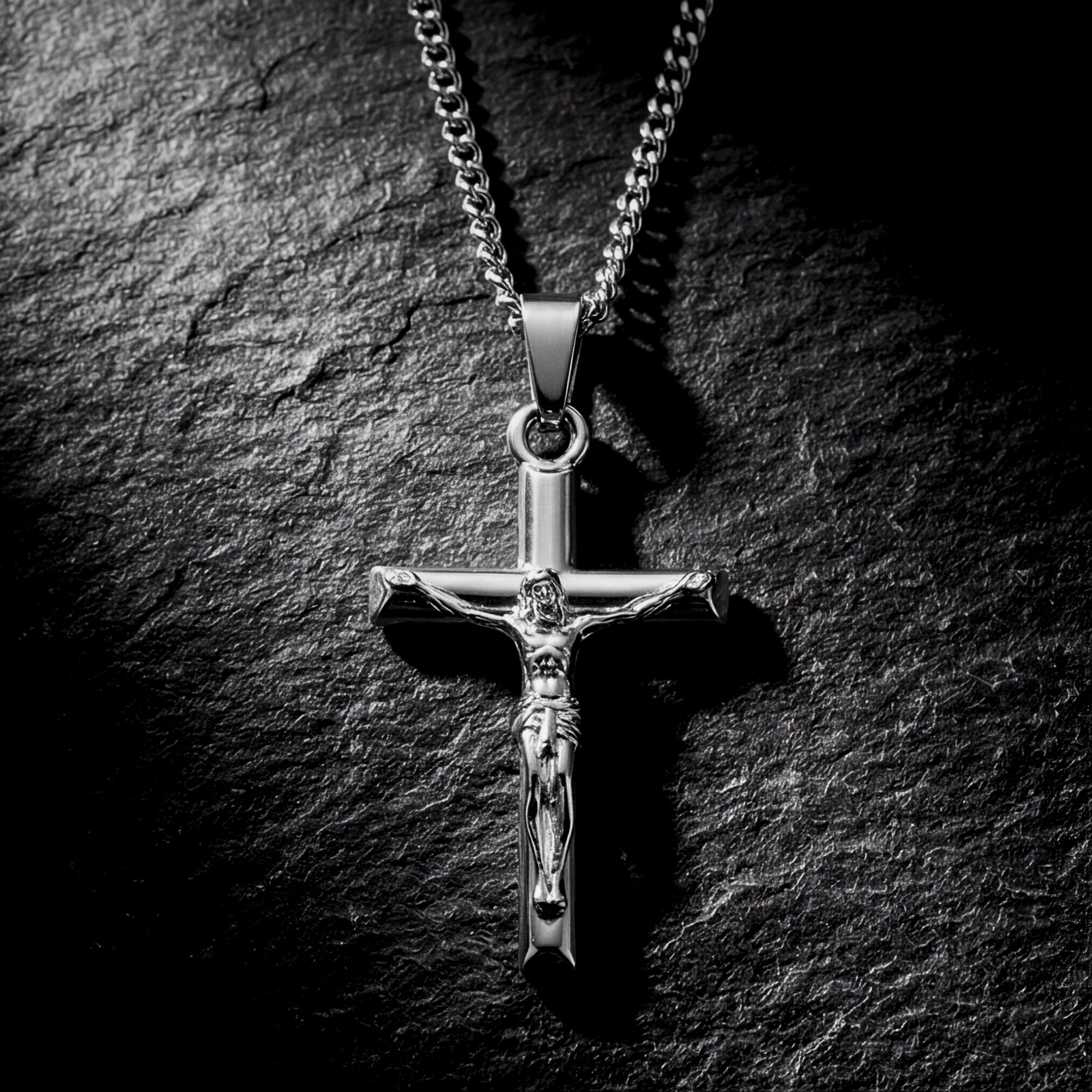 Silver stainless steel crucifix pendant necklace laid flat on a dark textured stone background.