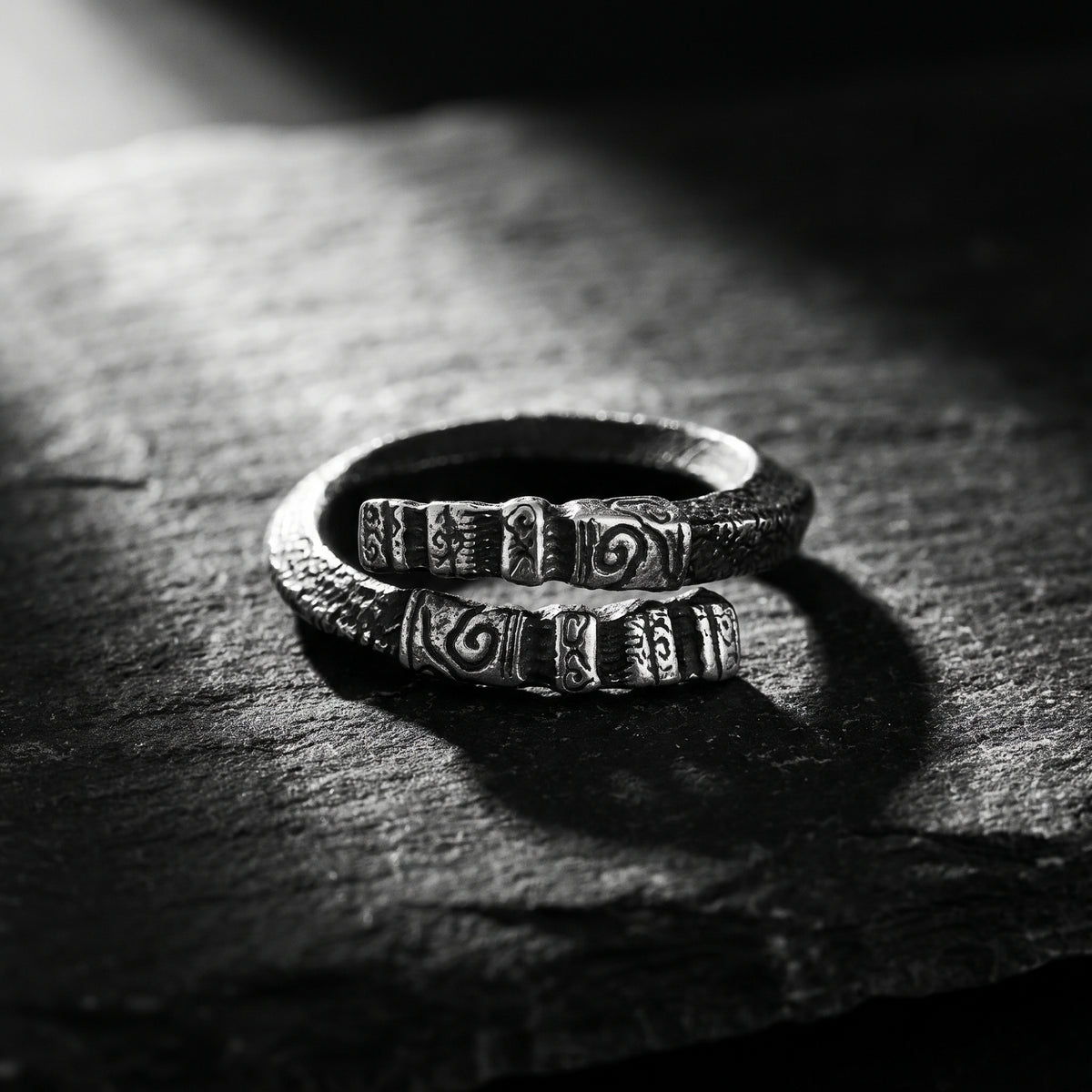 Carved silver open-band ring resting on dark stone, with its two open ends and spiral relief motifs visible.