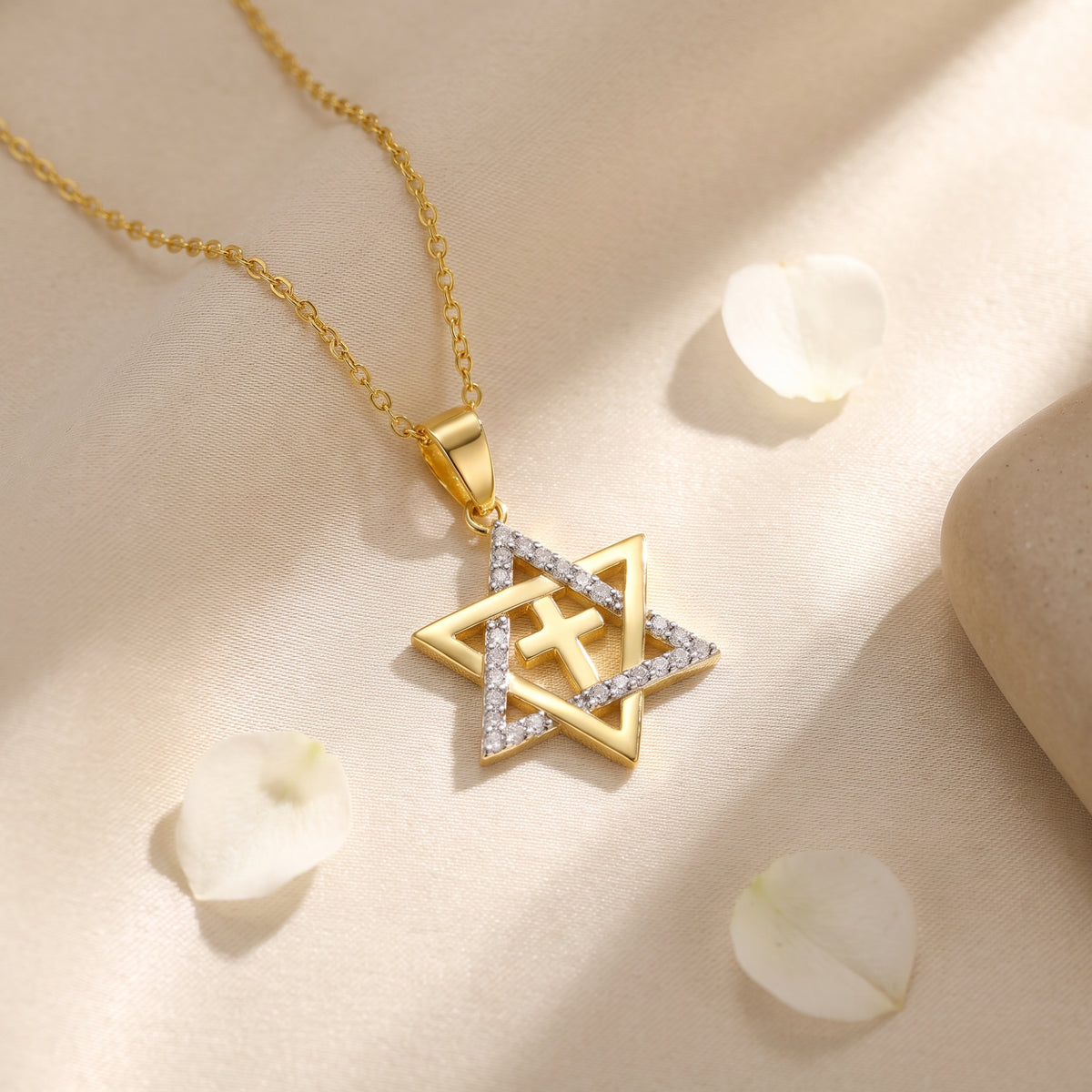 Gold-tone Star of David cross necklace lying on cream fabric with white flower petals.