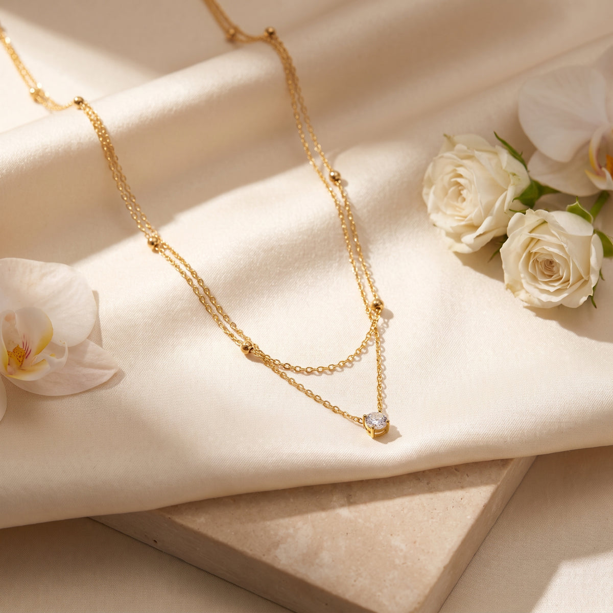 Angled view of the gold-tone layered necklace with a round clear pendant draped over satin among white flowers.