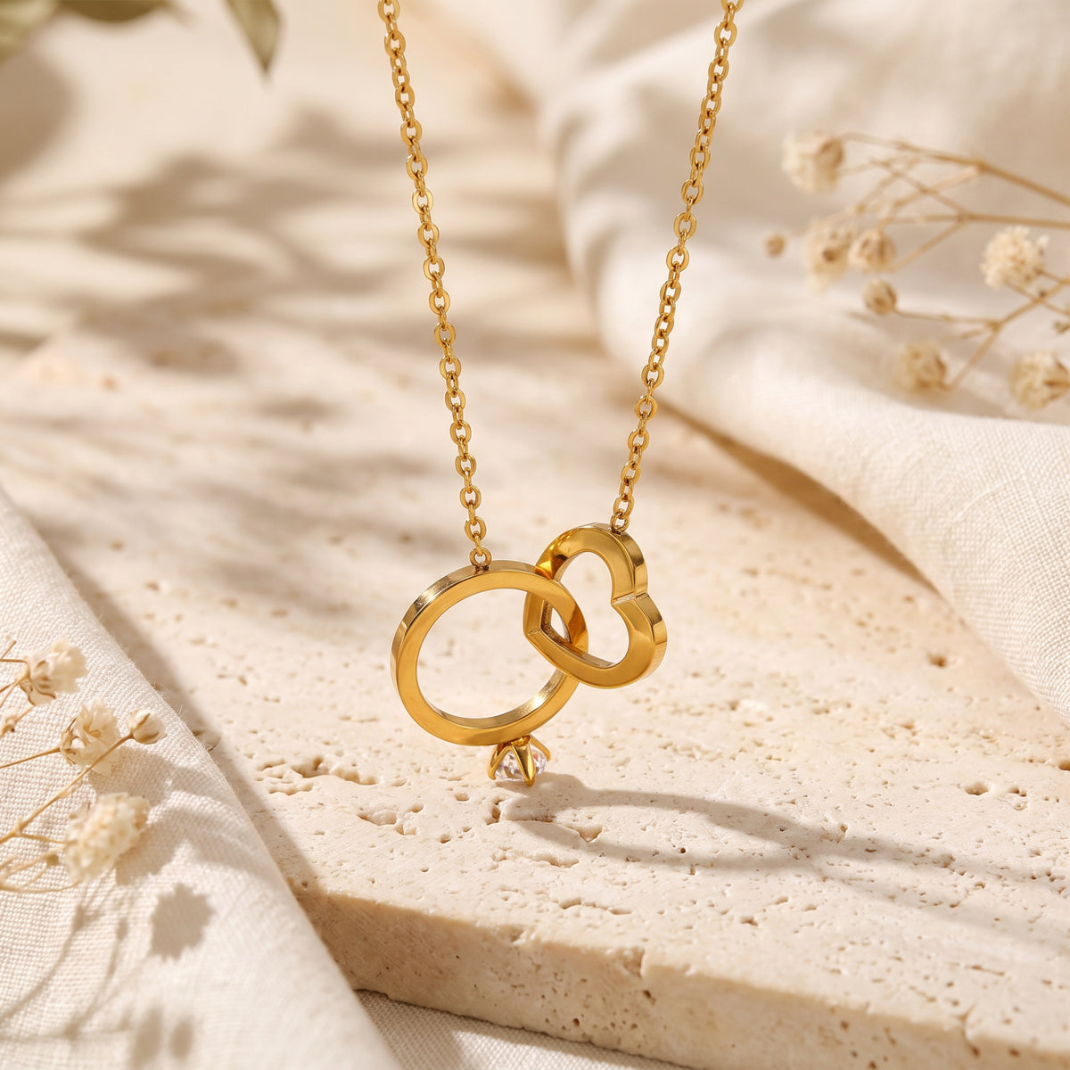 Upright gold-tone interlocking ring and heart pendant with a cubic zirconia accent suspended above beige stone.