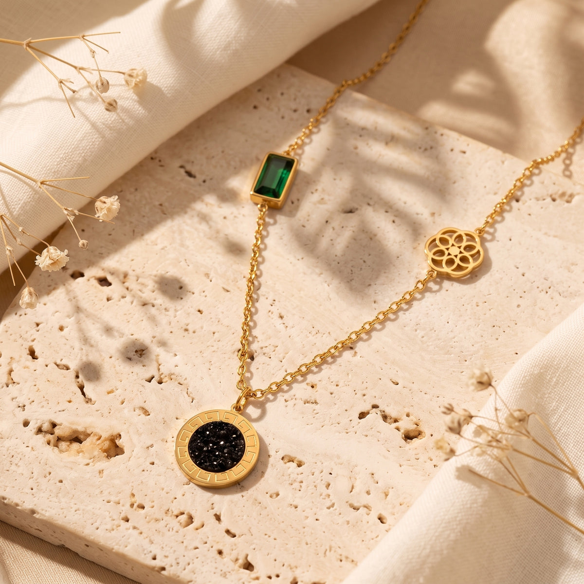 Gold-tone station necklace with a green crystal, floral wheel, and black medallion laid on textured stone.