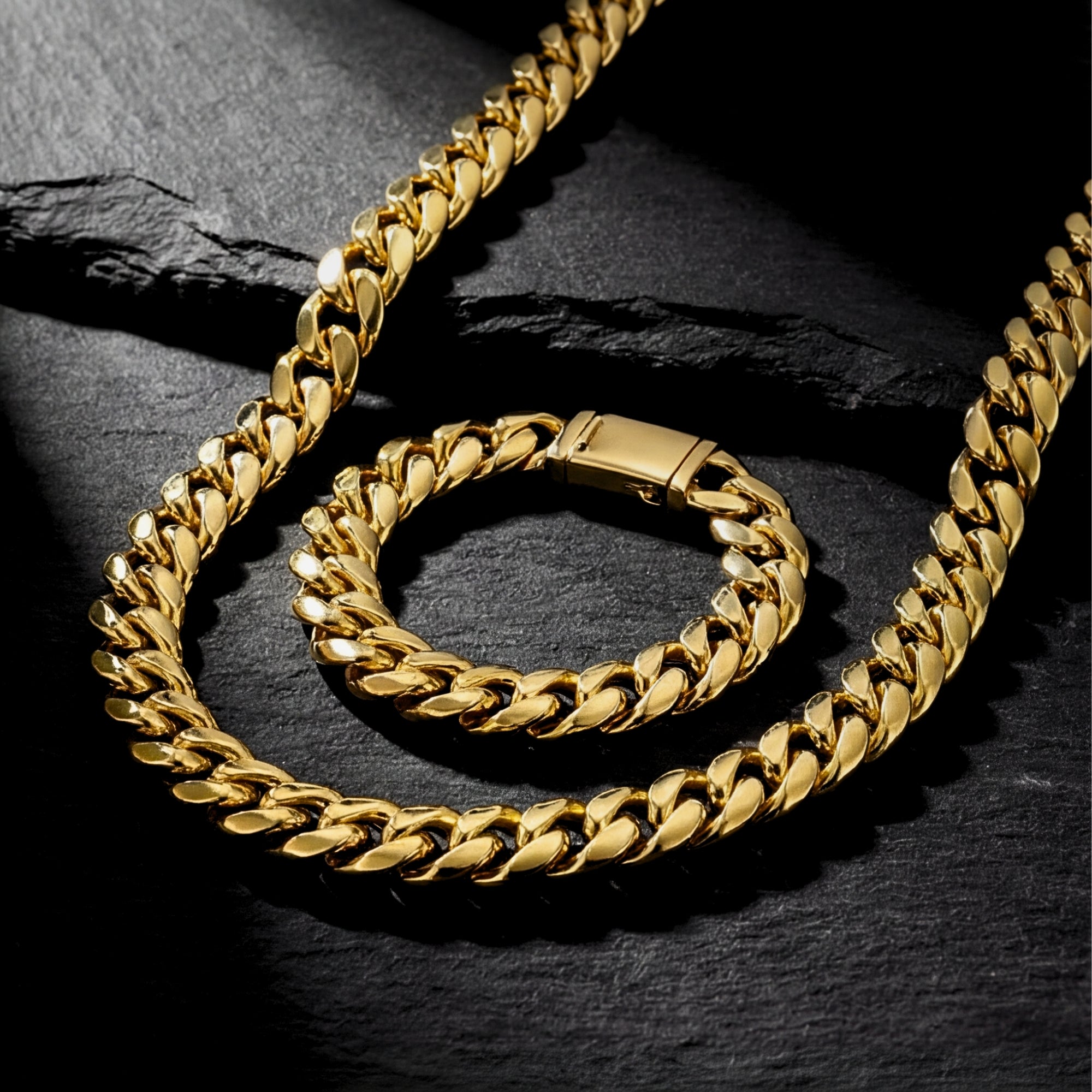 Angled view of a 10mm gold stainless steel Cuban link chain and bracelet set on a dark slate surface.