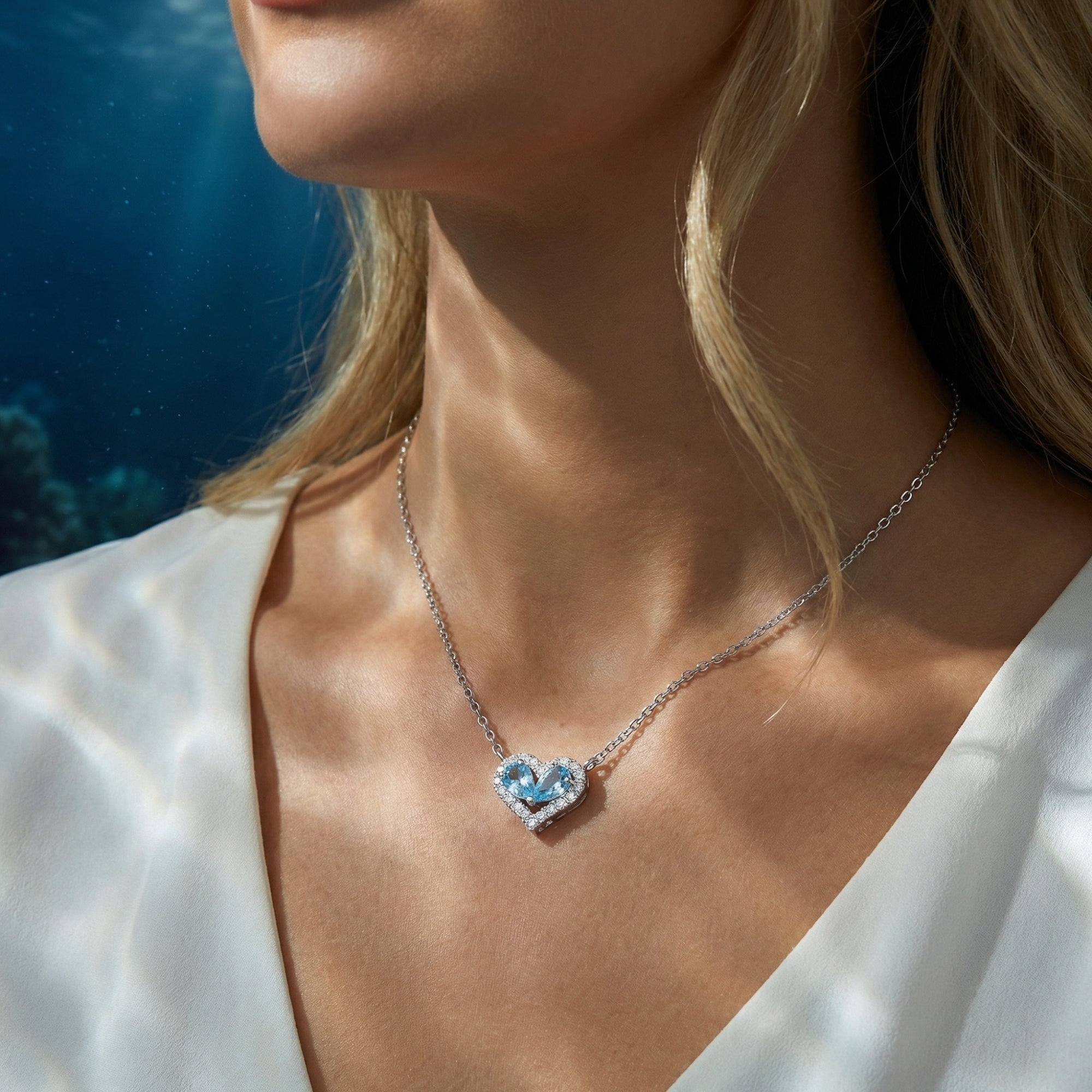 Model wearing the silver Maris Duo aquamarine heart necklace against a sunlit oceanic background.