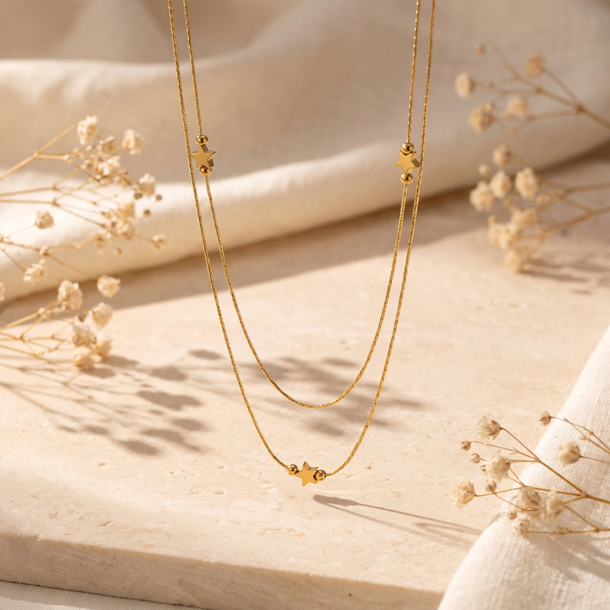 Gold-tone double-strand star necklace hanging above a beige stone block with dried flowers.