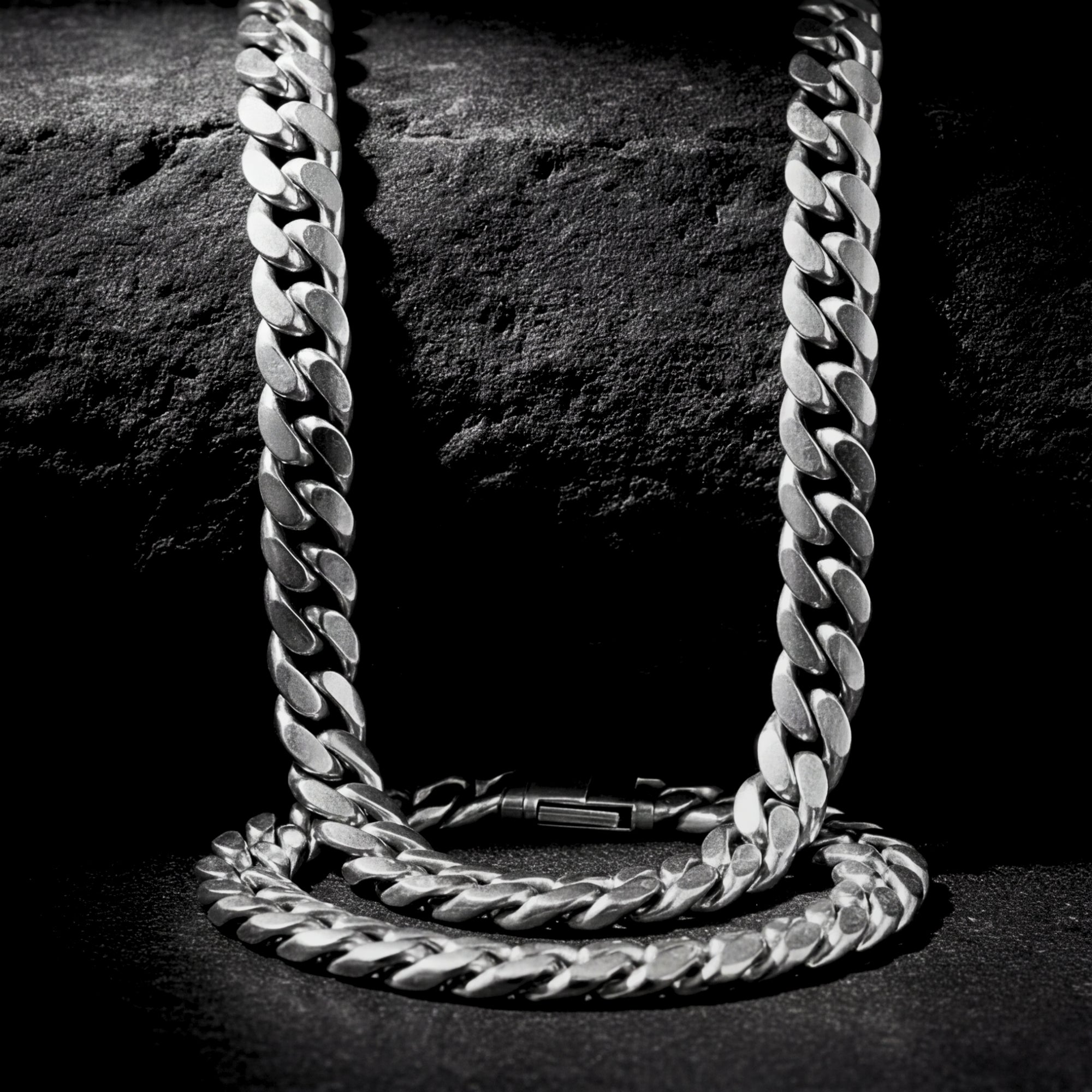 Close-up of the silver stainless steel Cuban link chain and bracelet set on a dark rock.
