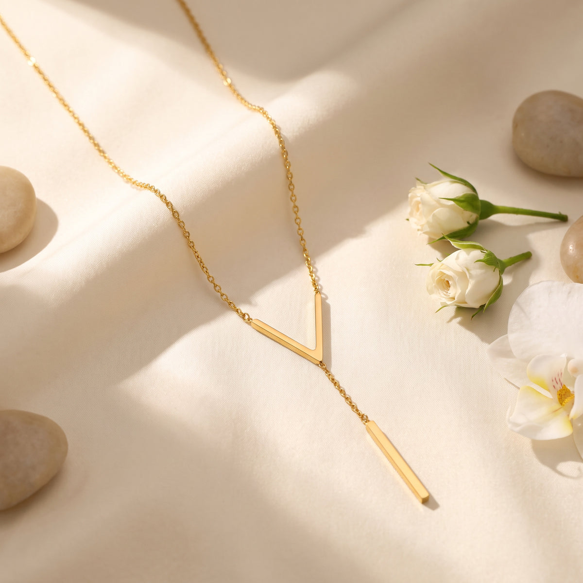 Gold-tone chevron lariat necklace arranged on draped cream fabric with white roses and smooth stones.