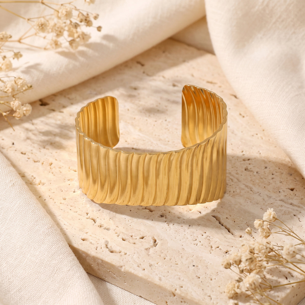 Upright front view of an open-back gold-tone ripple cuff bracelet on a stone surface.