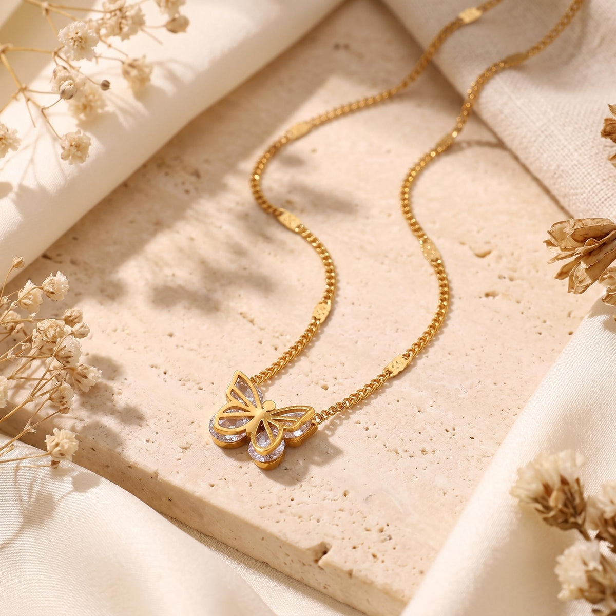 The gold-tone openwork butterfly necklace displayed flat on a stone tile with dried florals.