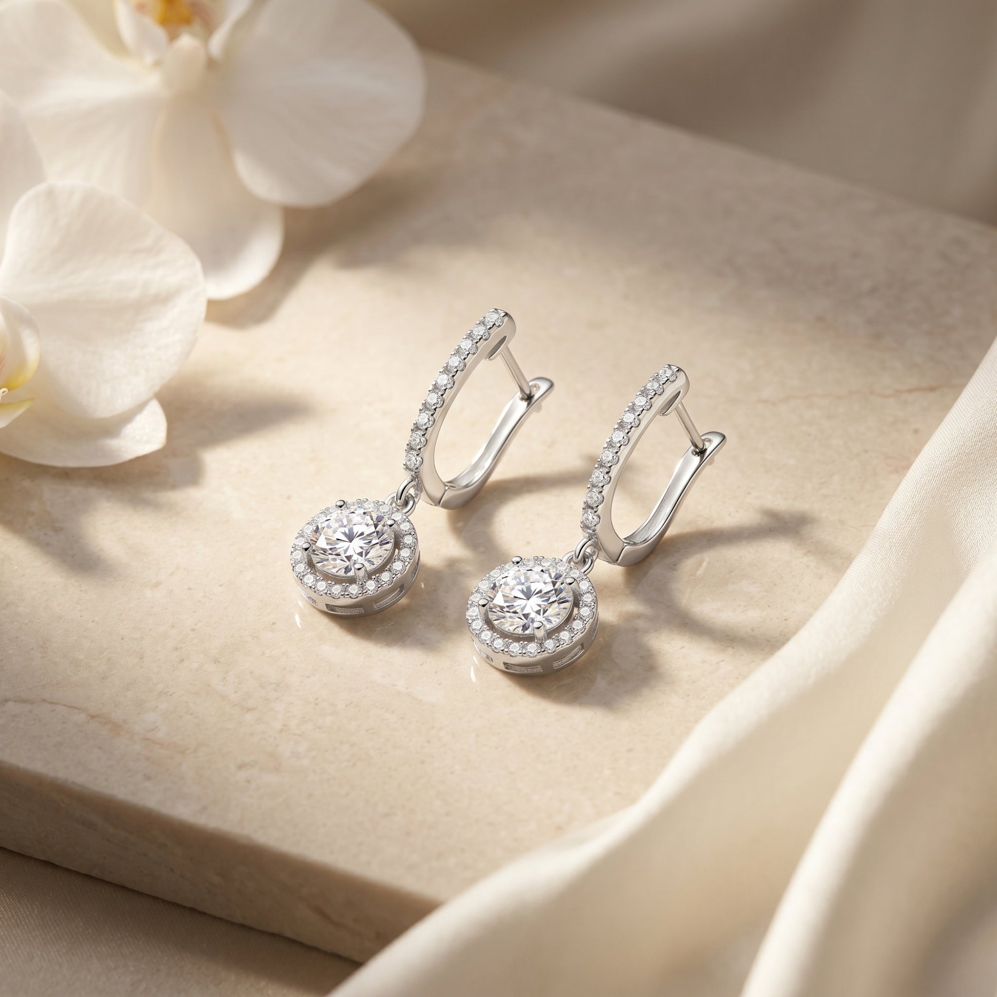 Pair of silver round moissanite drop earrings on a stone surface with white orchids.