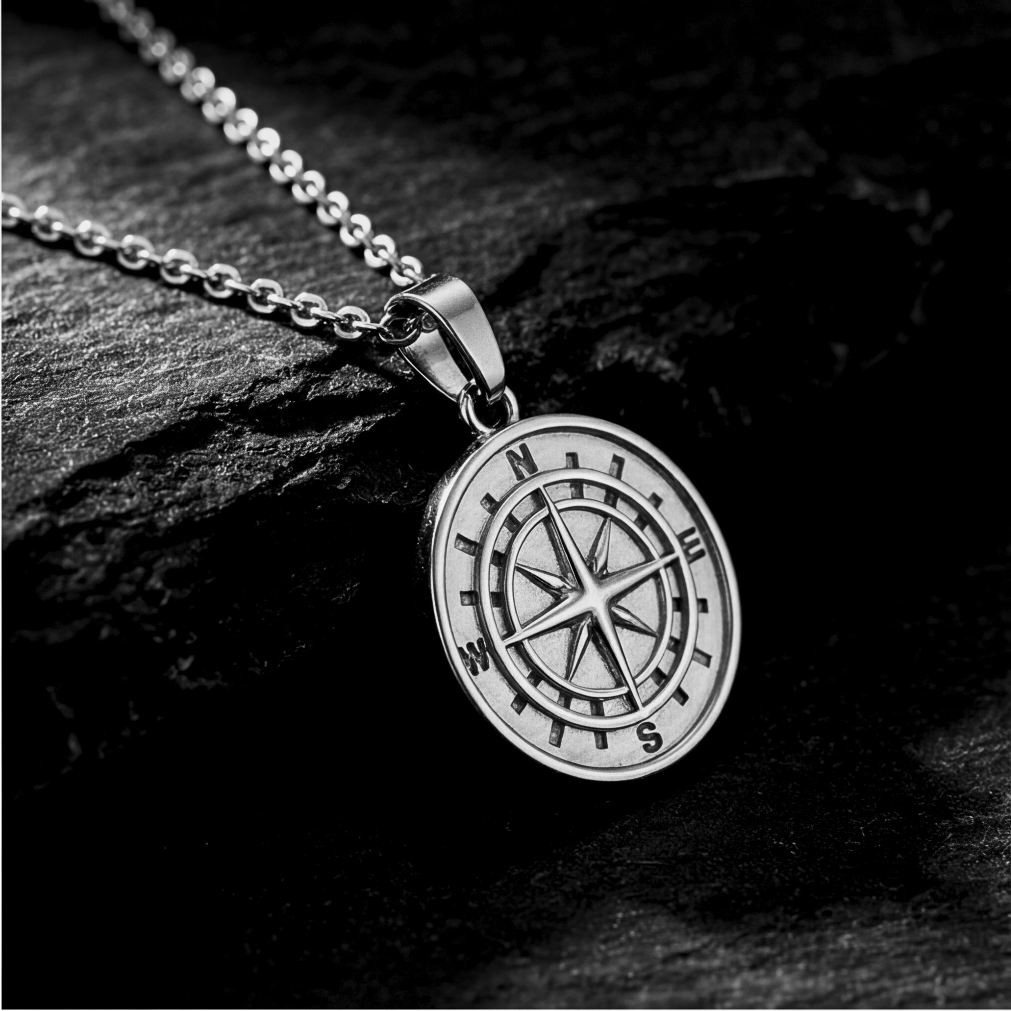Angled view of the silver stainless steel compass pendant on a chain, displayed on a dark stone background.