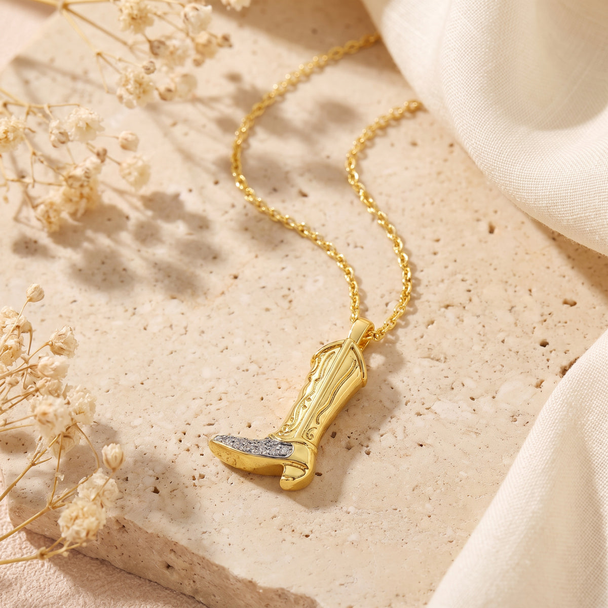 Gold-tone cowboy boot pendant necklace resting flat on a beige textured tray alongside dried flowers.
