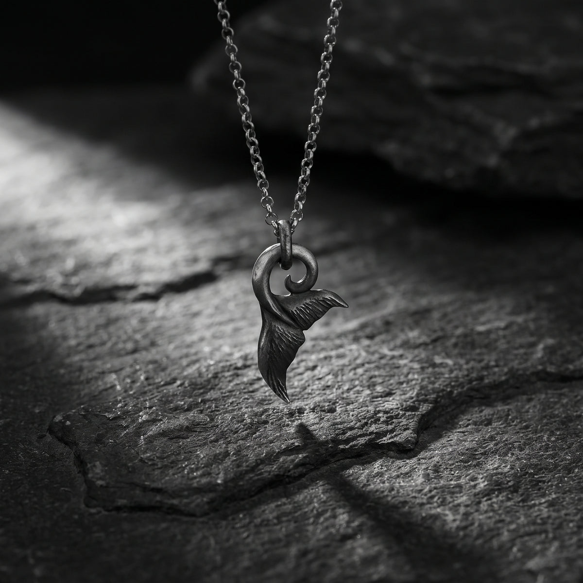 Front view of the silver whale tail fluke pendant hanging from its chain above dark slate.
