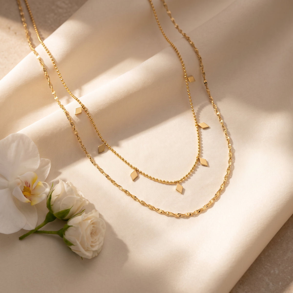 Gold-tone layered station necklace arranged on cream fabric beside white flowers.