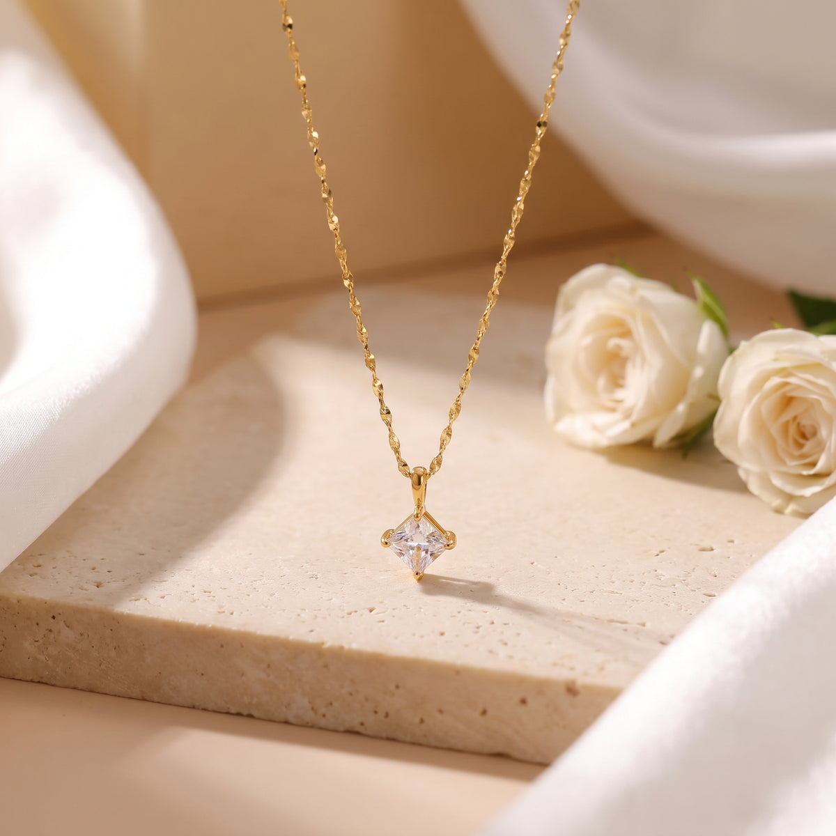 Gold-tone square solitaire pendant necklace hanging over a beige stone slab beside white roses.