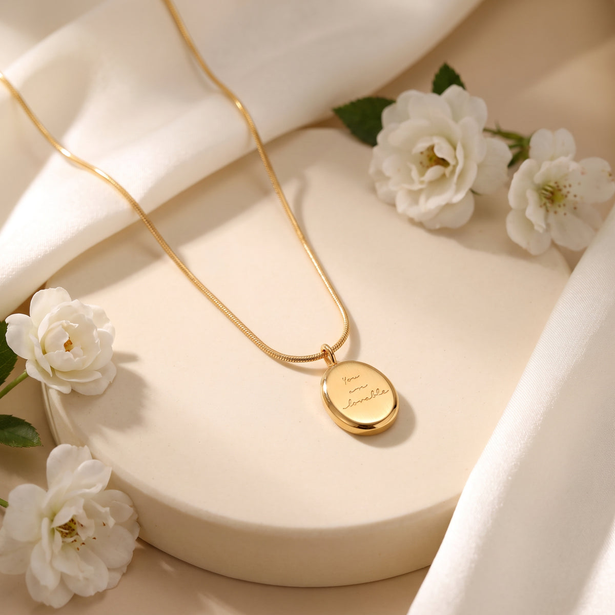 Gold-tone engraved oval medallion necklace displayed on a round cream pedestal with white flowers.