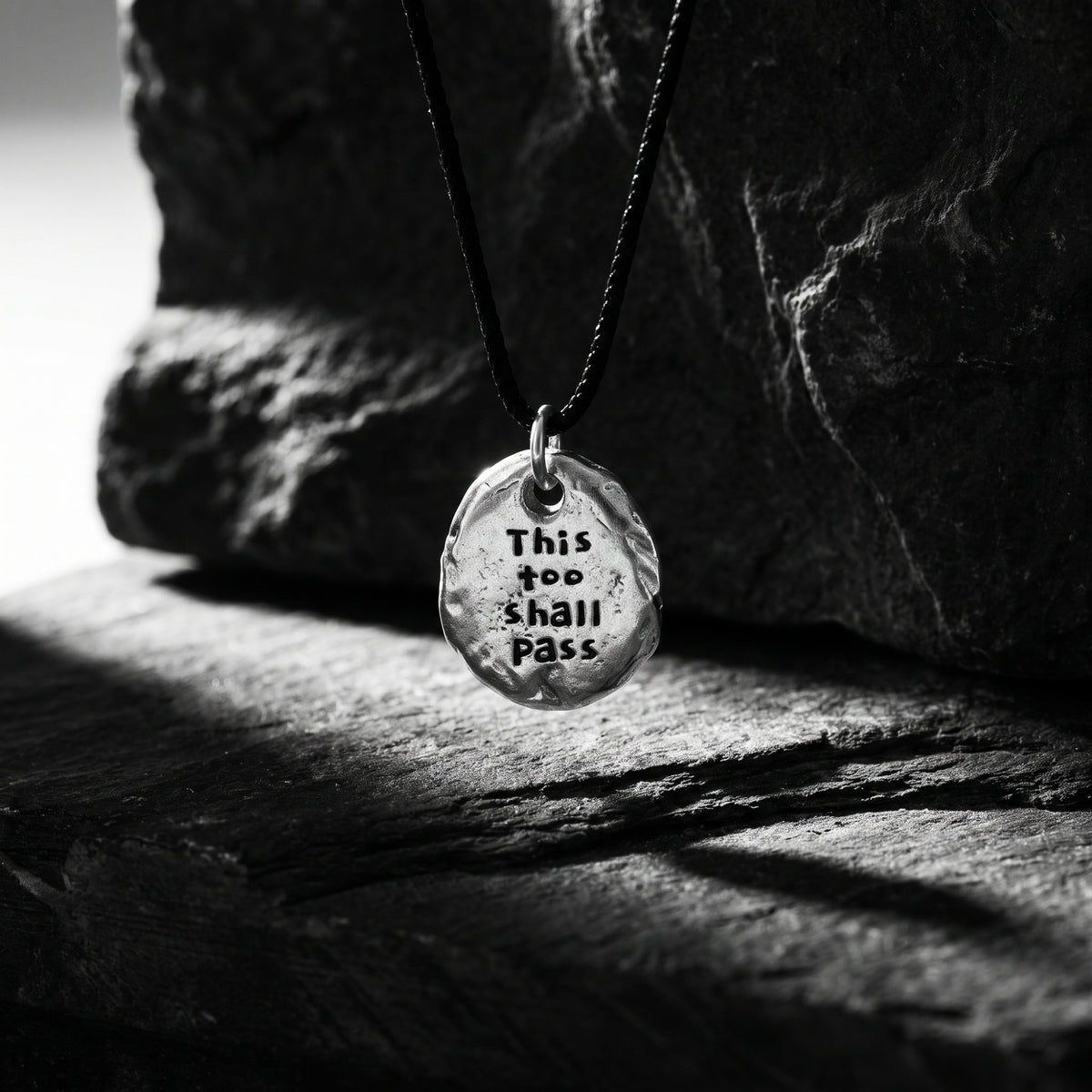 Engraved silver pebble pendant hanging from a black cord in front of dark slate rocks.