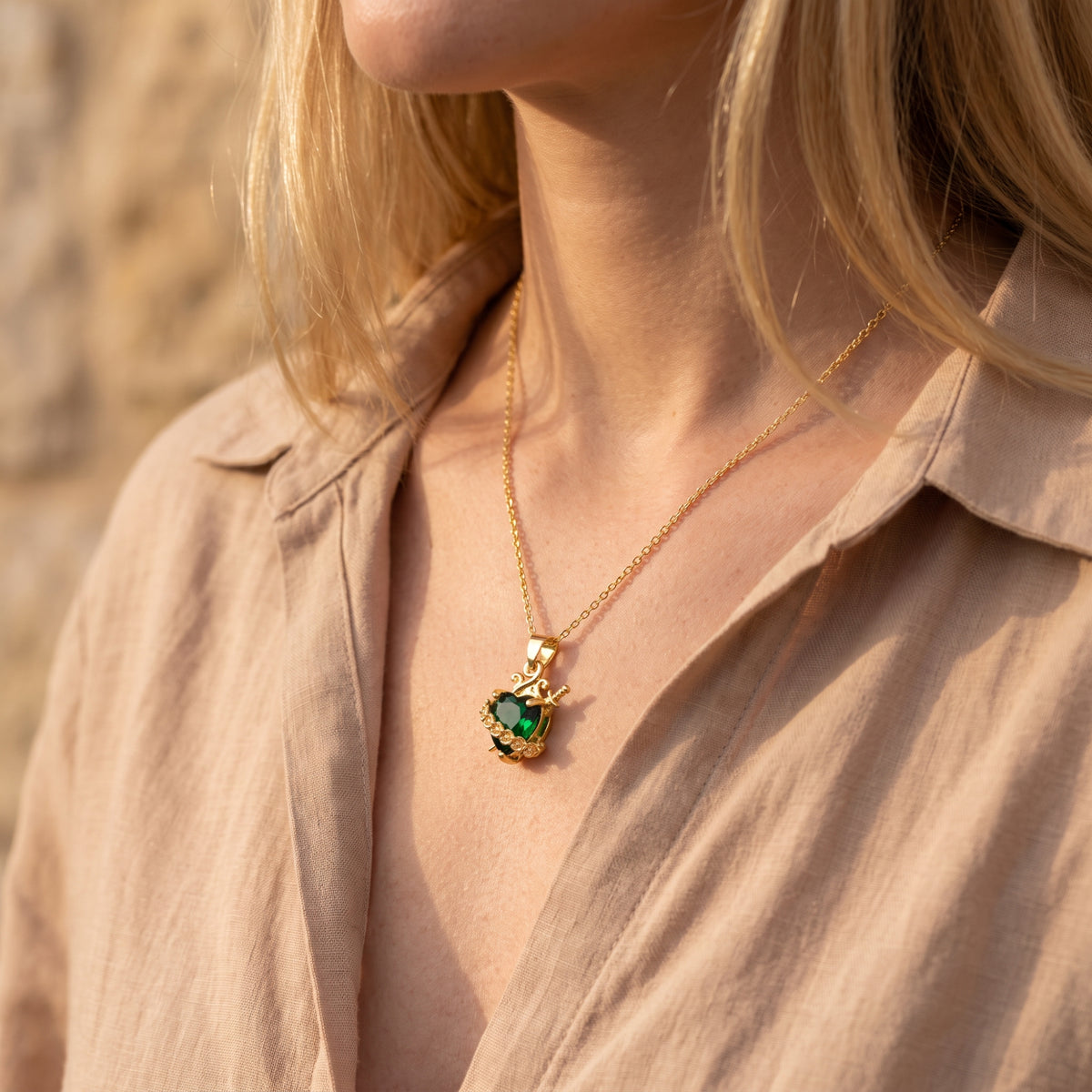 Model wearing the gold heart and dagger necklace with an open beige shirt in warm sunlight.