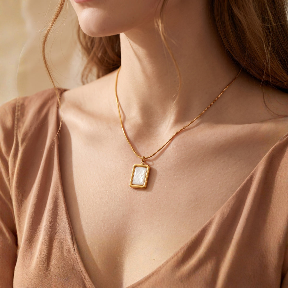 Woman wearing a gold-tone rectangular pendant necklace with a quilted white inlay over a low V-neck tan blouse.