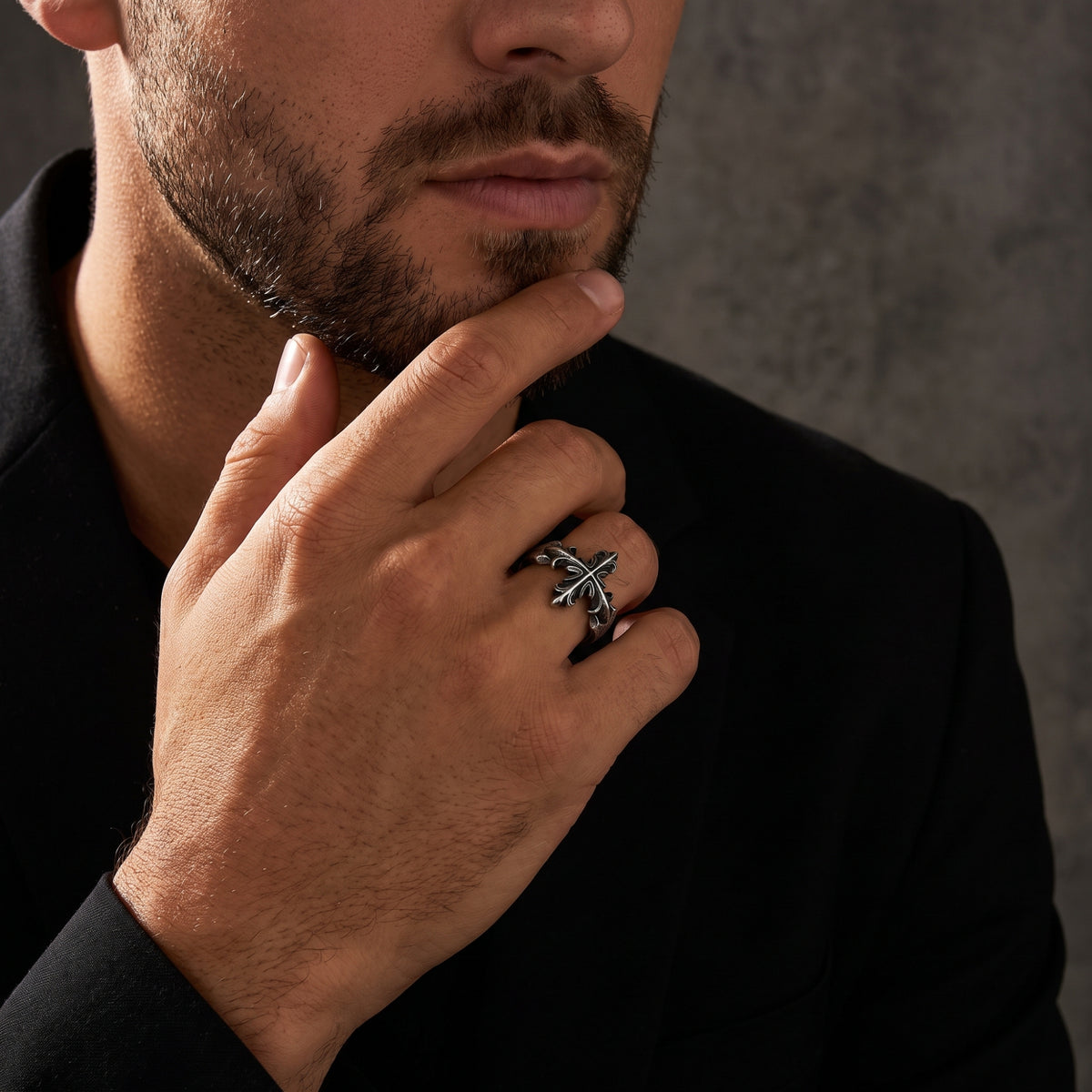 Wider portrait of a man wearing the silver Gothic cross ring while touching his chin in a black jacket.