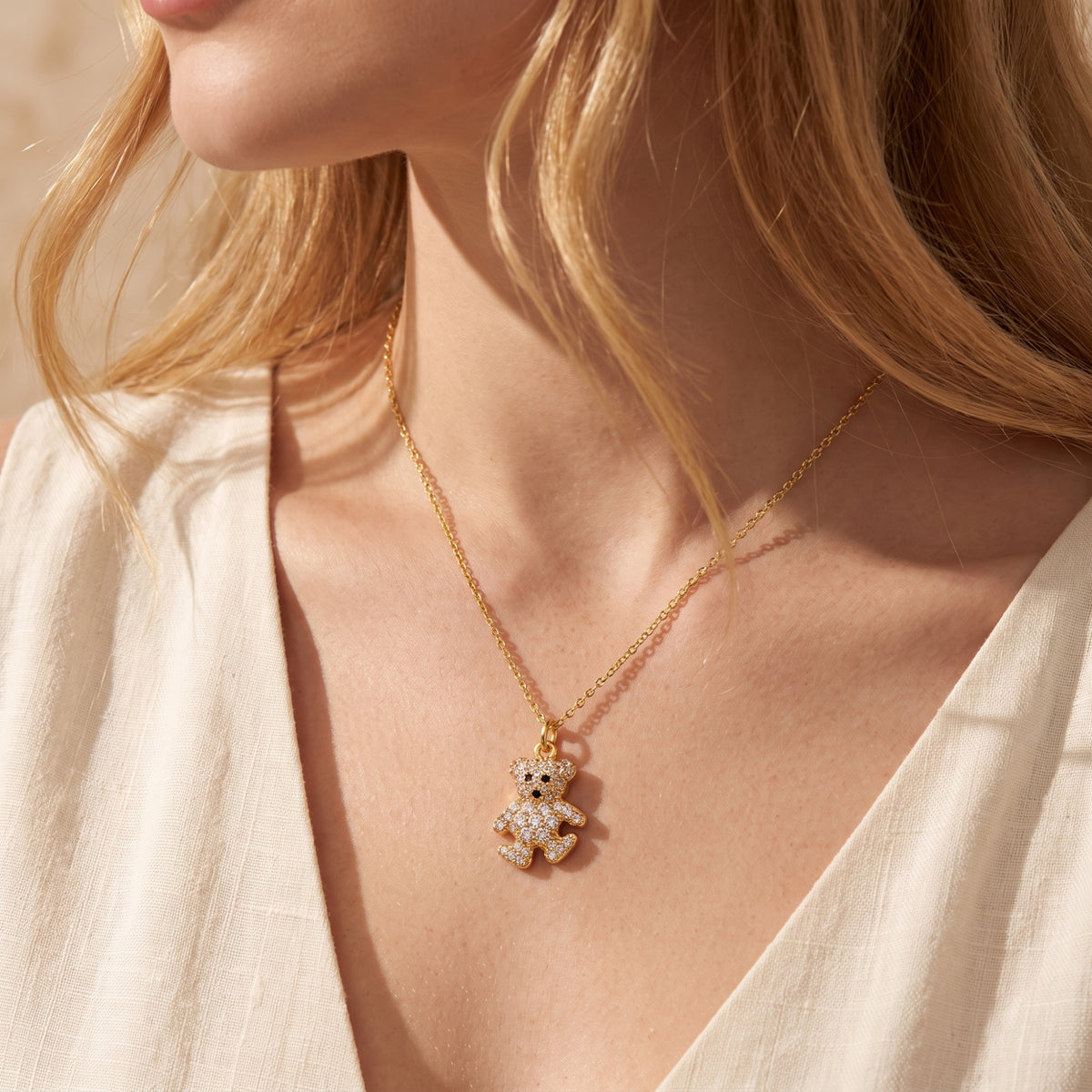 Close-up of a woman wearing a gold-tone crystal teddy bear necklace over a cream top.