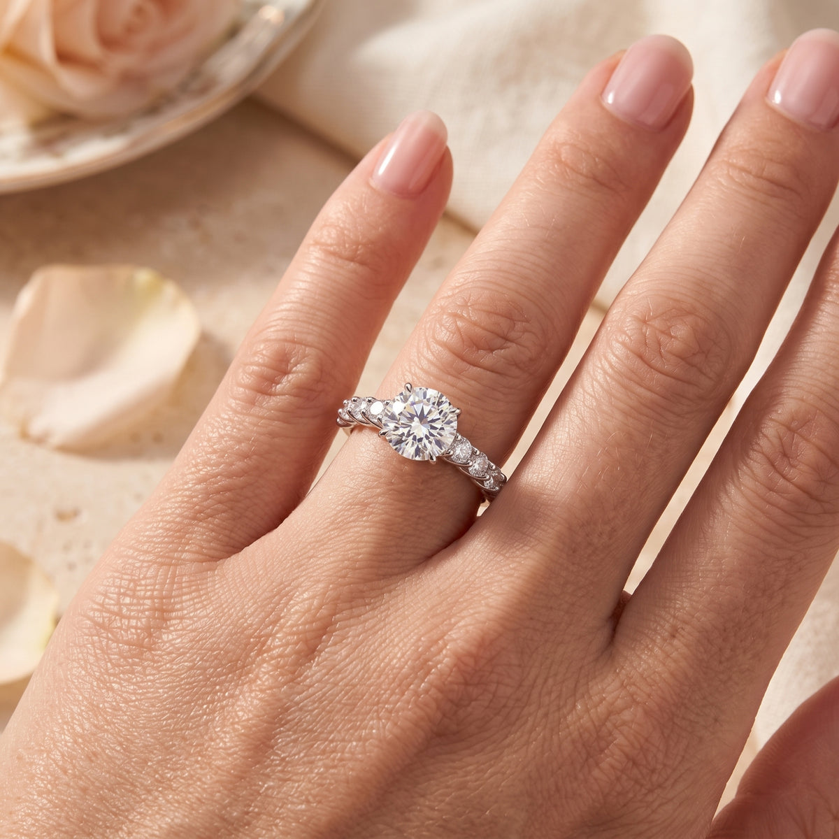 Silver round-cut solitaire ring with accent stones worn on a hand above light-colored flower petals.