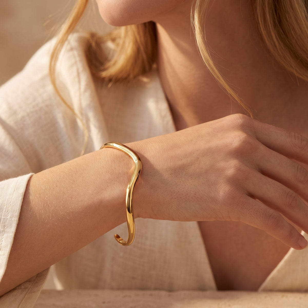 Close-up of a model's hand and wrist wearing the gold-tone open wave cuff beside a white sleeve.