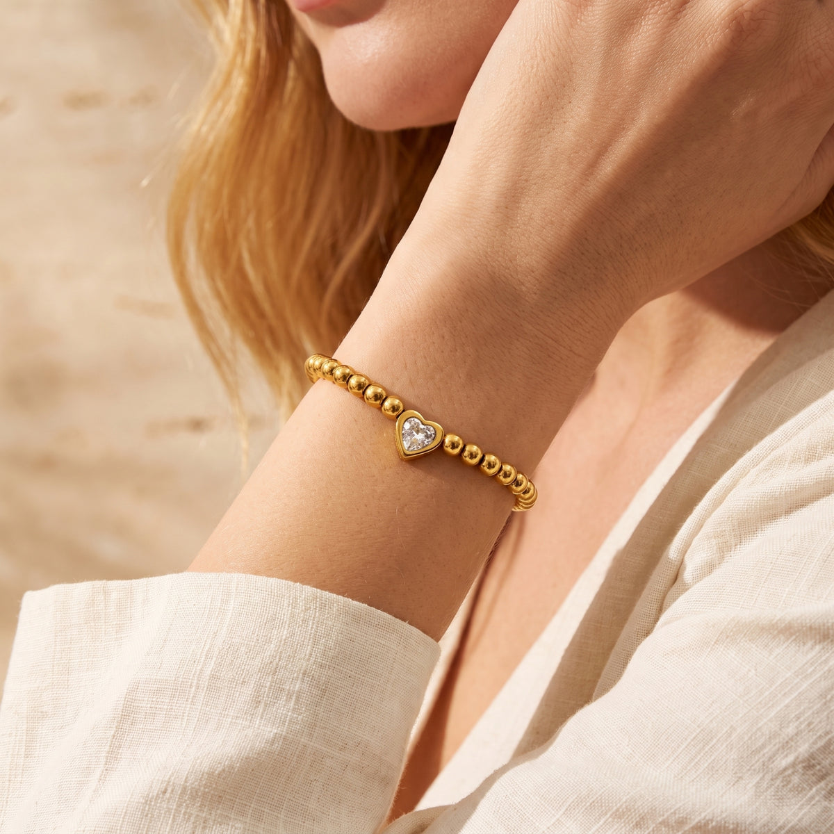 Close-up of a gold-tone beaded bracelet with a heart-shaped clear stone worn on a wrist beside a white sleeve.